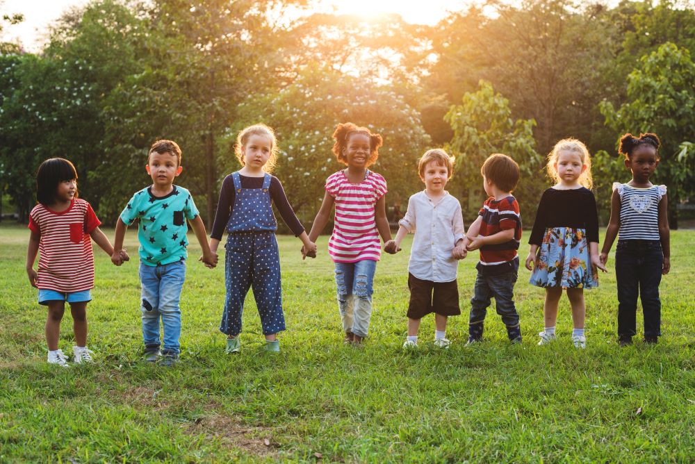 8 children of various ages and ethnicities holding hands as they stand side by side outdoors while the sun shines brightly behind them.