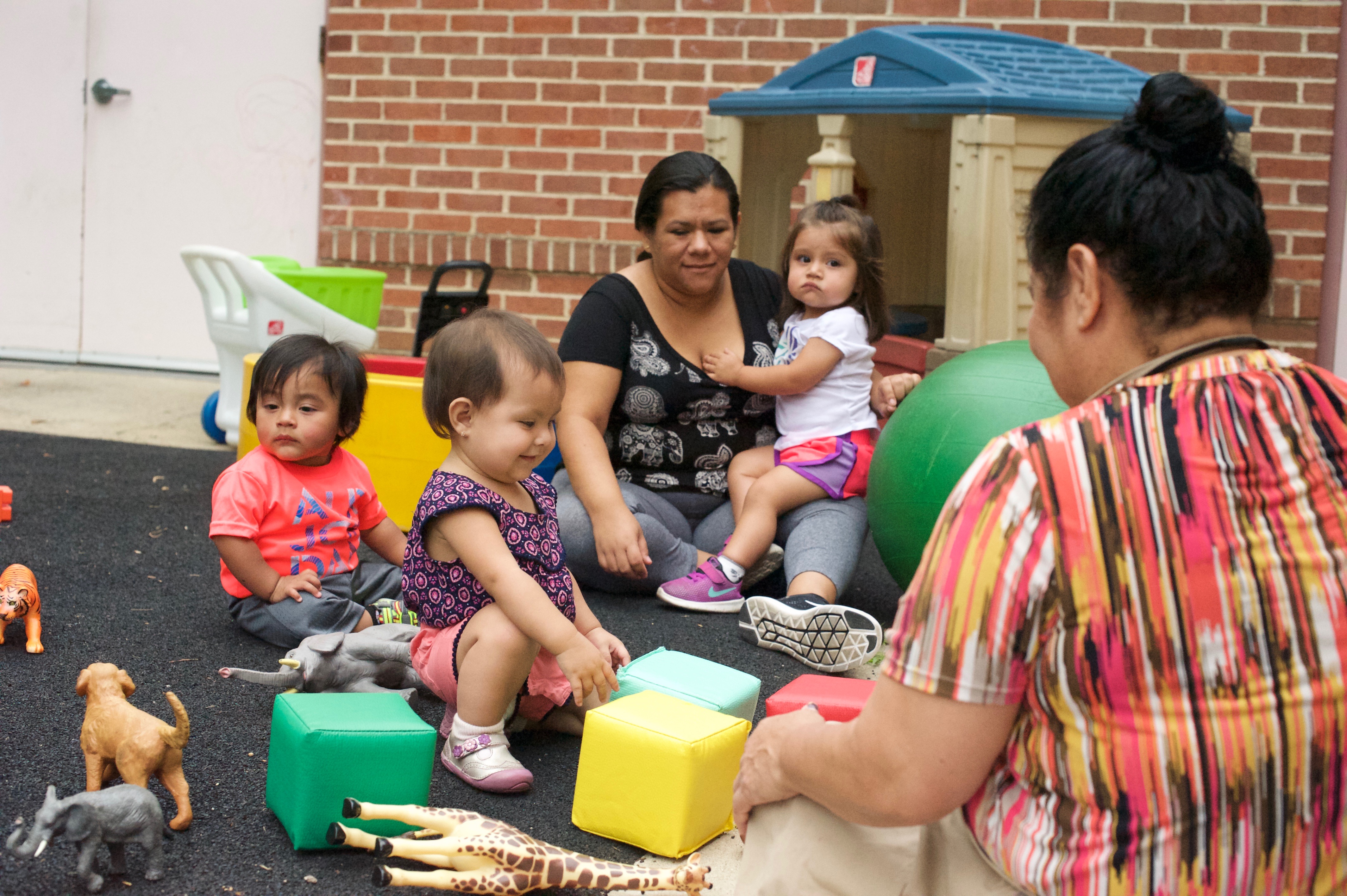 Two adults supervising several small children playing on the floor with toys.