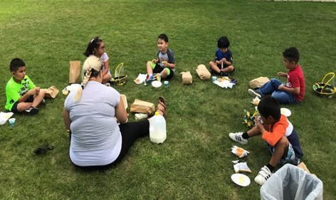 Niños almorzando afuera, acompañados por una maestra