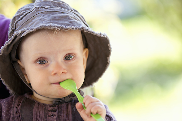 Baby wearing a floppy hat with a green spoon in its mouth.