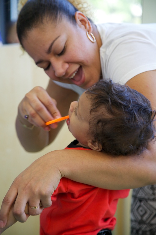 Teacher brushing baby's teeth while holding child close to her side.