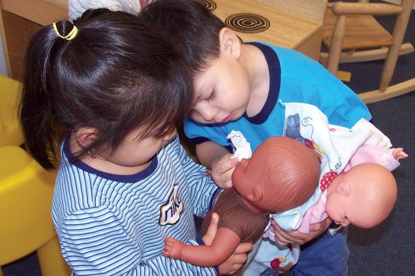 A boy and girl pretending to wipe away tears from a doll's eyes.