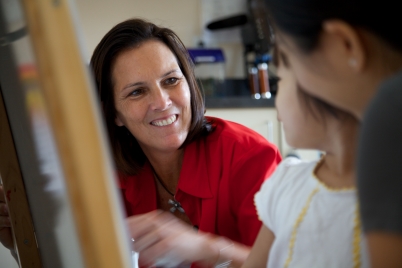 Adult smiling at two young children as they draw.