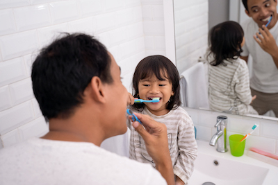 Father standing while young daughter is sitting on sink both brushing their teeth.