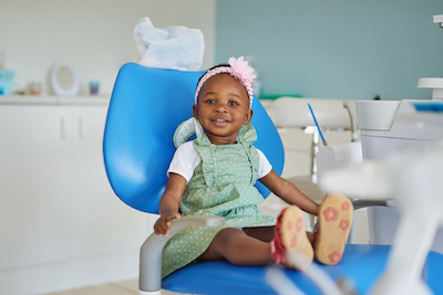 Una niña pequeña con un lazo rosa en la cabeza, sentada en el sillón del dentista lista para un examen.