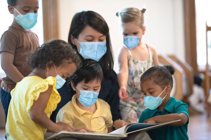 A group of multi-ethnic children wearing face masks gathered around a teacher who is reading a children's book to them.