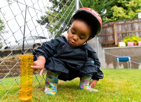 Young boy reading a rain gauge placed against a fence in a playground.