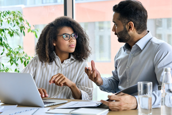 A woman and man having a discussion while looking at a laptop screen and taking notes.