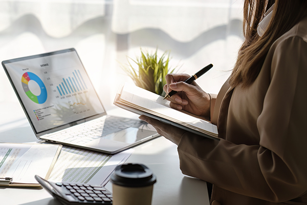 Woman making calculations using pen and paper while watching graphics from a laptop.