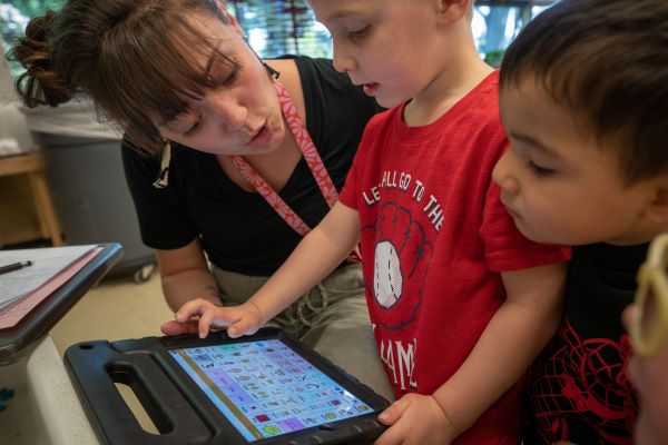 Teacher assisting a child to use a didactic application in a tablet, while another child watches.