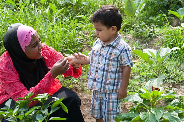 Teacher talking with a child about a vegetable, when being outside surrounded by plants and weeds.