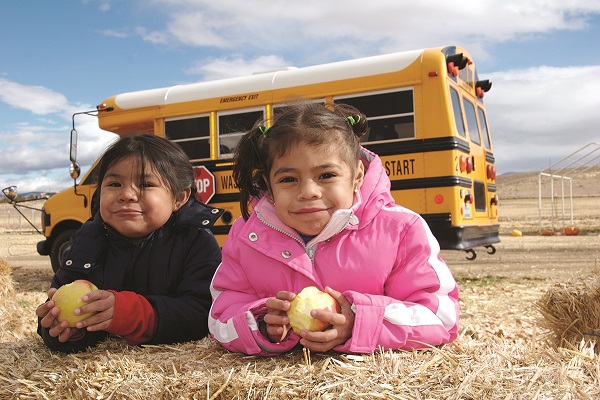 Two children laying on the ground outside, while a school bus is parked in the background.