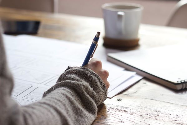 Close up of a person writing in a notebook with a cup of coffee in the backgroud.