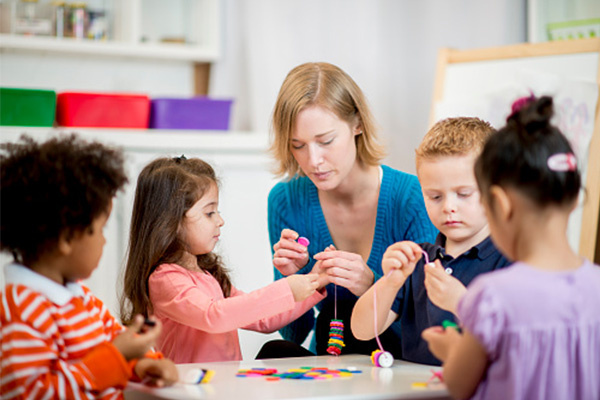 Teacher or volunteer helping children to make a necklace with beads.