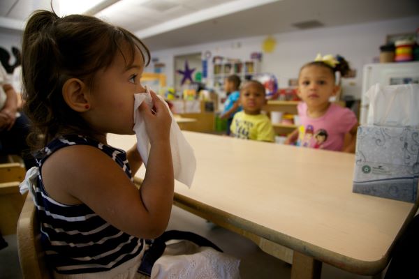 Niña limpiándose la nariz con un pañuelo de papel mientras está sentada en una mesa en su salón de clases.