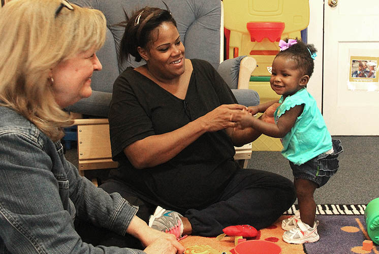 a baby on a play rug smiles at two adults helping her stand