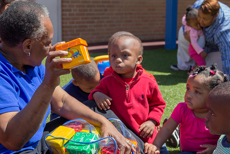 A teacher demonstrates a block to a group of children outside