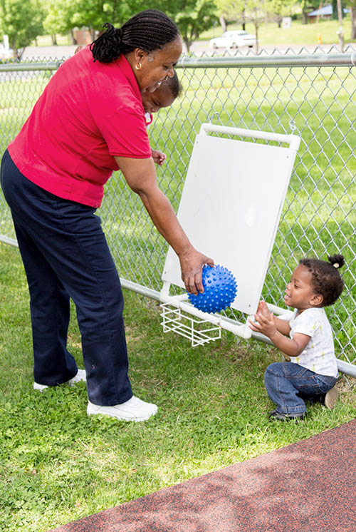 a teacher holding a baby hands a ball to a child sitting on the grass