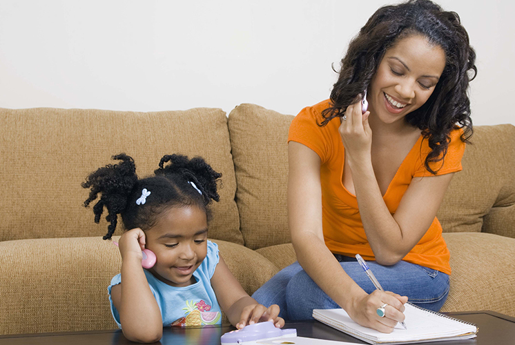 A mom on the phone takes notes while daughter plays nearby