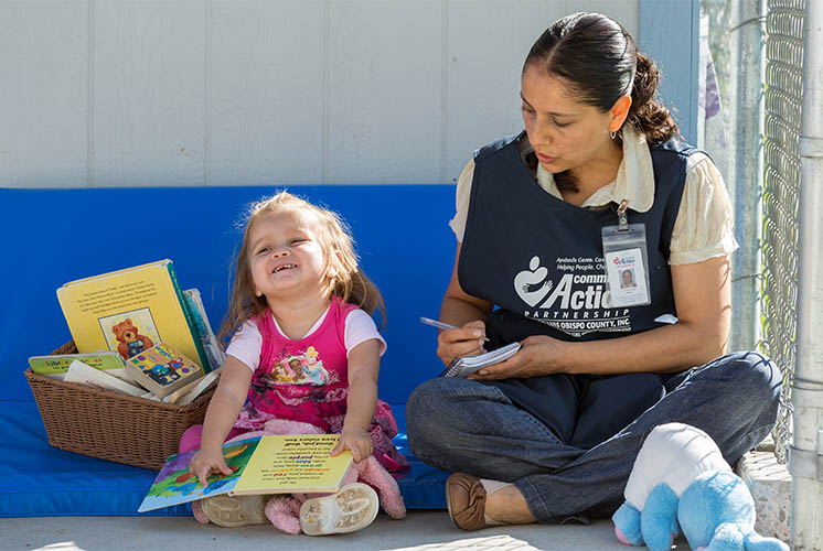 A caregiver sits on the ground near a child and takes notes