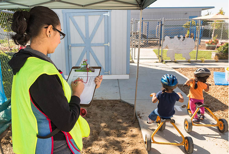 a careworker writes on a checklist as children ride by on tricycles outdoors