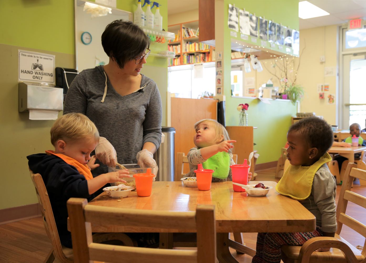 Maestra preparando alimentos para unos niños sentados a la mesa.