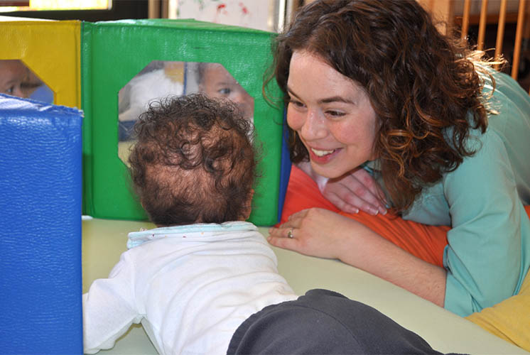 A toddler lies on the floor in front of play mirrors with a teacher looking on