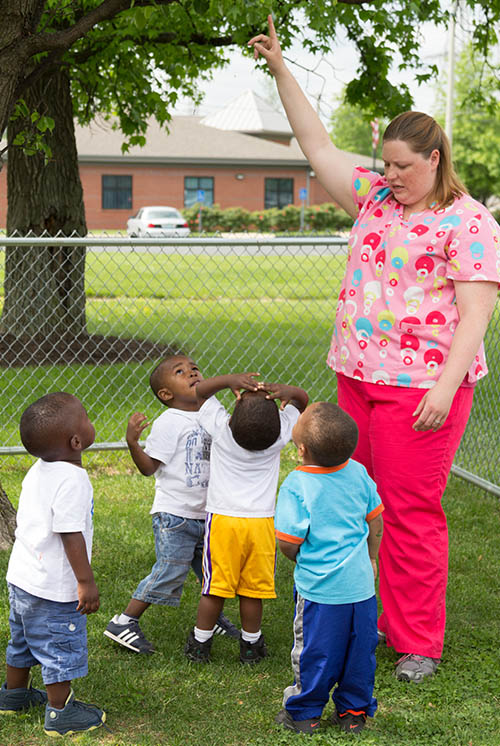 a teacher and group of kids stand under a tree looking up, teacher pointing up at tree branches